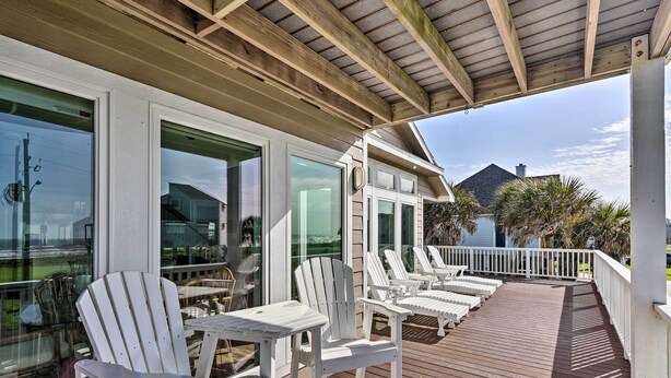 Une terrasse extérieure couverte, dotée de chaises et de tables Adirondack blanches, qui offre une vue sur un plan d’eau et des palmiers.
