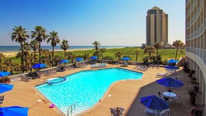 A resort pool area with lounge chairs and umbrellas, overlooking a beach and a tall building. - Heated Pool + Gulf View: Galvestonian Resort Condo (Galveston)
