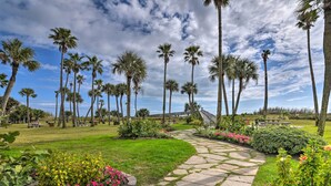 A pathway lined with palm trees and flowers, leading to a grassy area with benches and a picnic table. - Heated Pool + Gulf View: Galvestonian Resort Condo (Galveston)