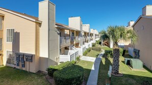 A row of single-story buildings with balconies, a well-maintained lawn, and a palm tree. - Balcony & Pool Access: Condo in Galveston! (Galveston)