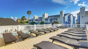 A deck with sun loungers and a pool, surrounded by blue buildings with white railings. - Balcony & Pool Access: Condo in Galveston! (Galveston)