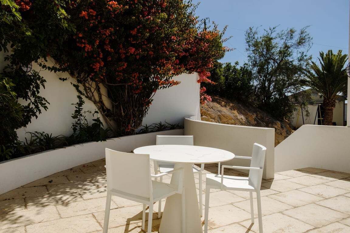 A patio with a round table and four white chairs, surrounded by a low white wall and various plants.