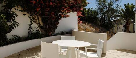 A patio with a round table and four white chairs, surrounded by a low white wall and various plants.