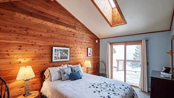 A cozy bedroom with wooden walls, a large bed with a floral bedspread, two bedside lamps, and a window with a view of the outdoors.