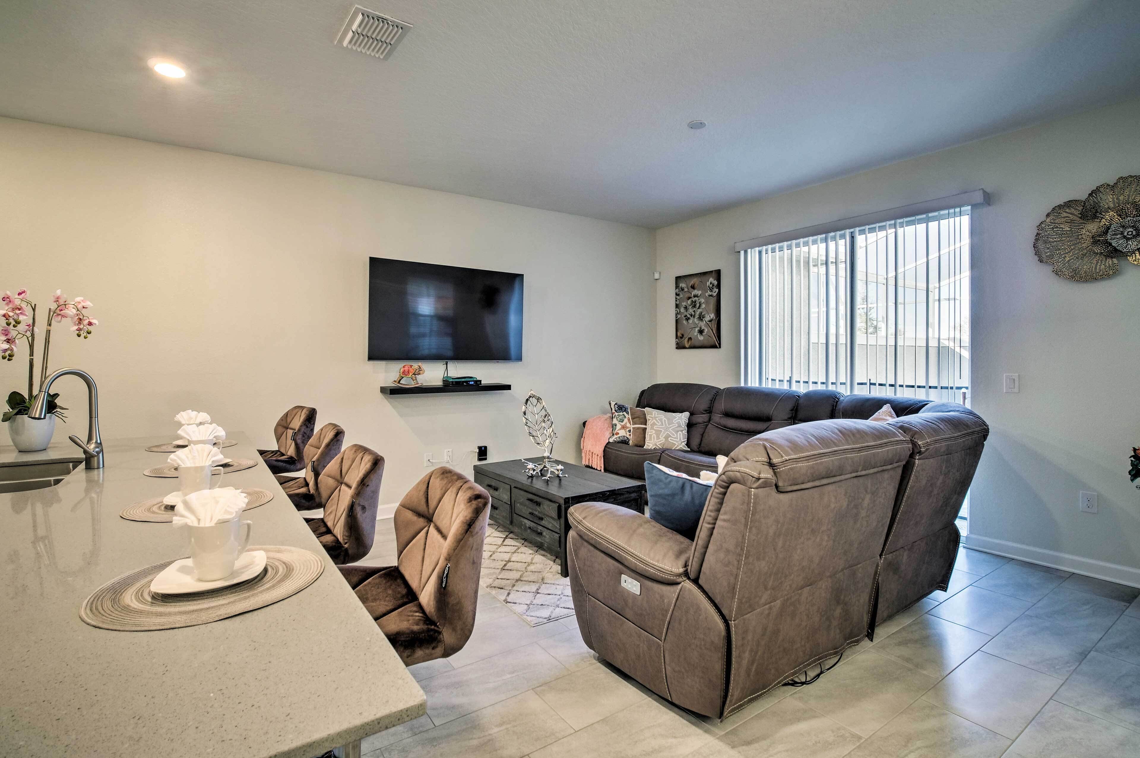 A modern living room with a sofa, a TV mounted on the wall, and a dining area with a table and chairs.