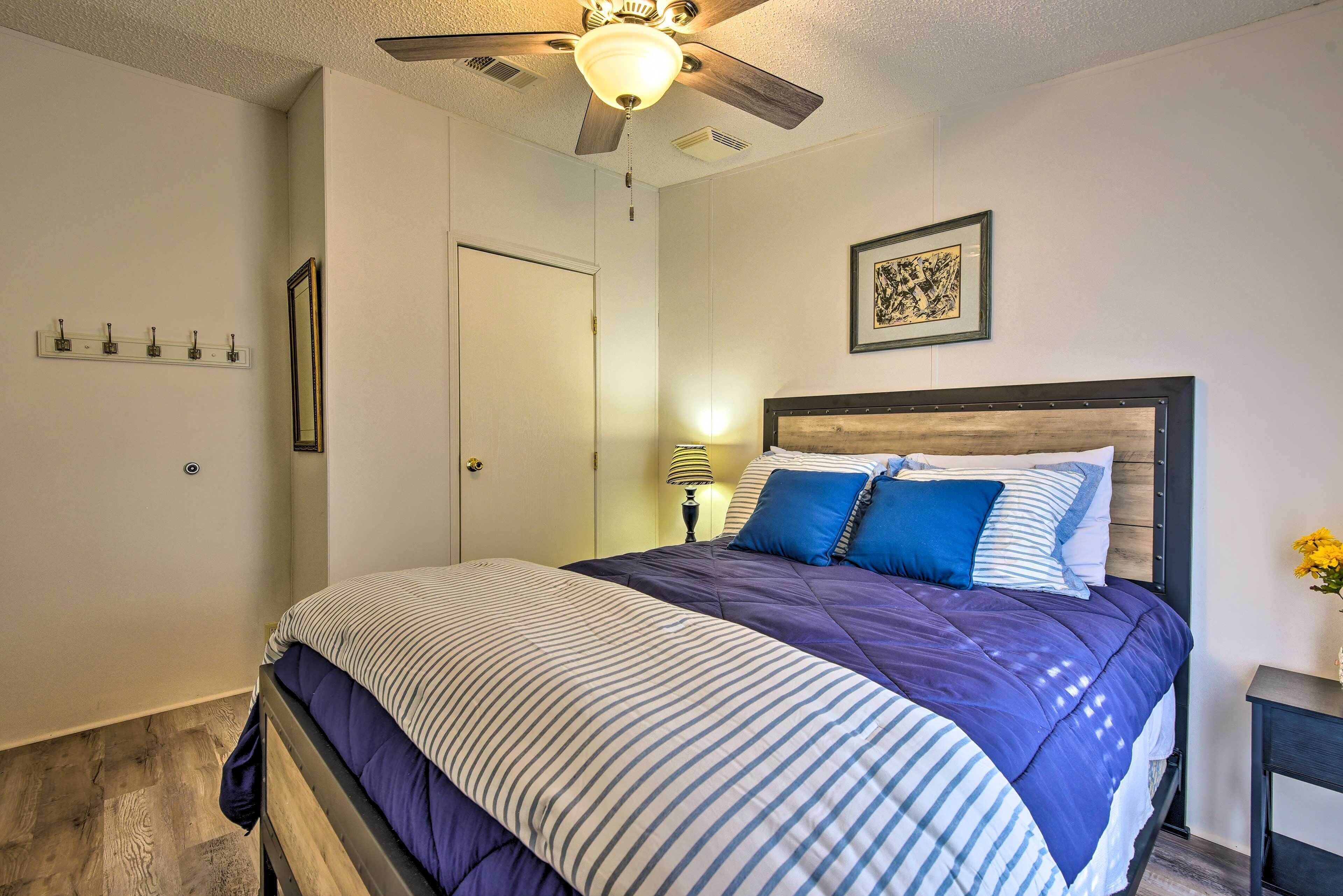 A neatly made bed with a striped comforter and blue pillows, a wooden headboard, a ceiling fan, and a bedside lamp.