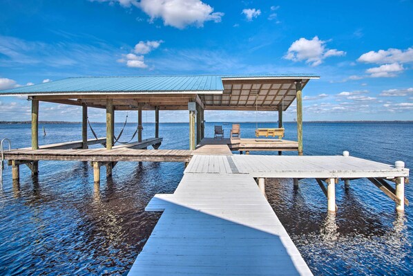 A wooden pier leading to a covered seating area with a bench and a table, extending over calm water.
