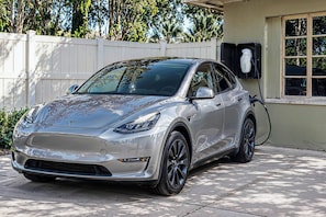 Un Tesla Model 3 plateado estacionado en una entrada con una cerca blanca y árboles al fondo.