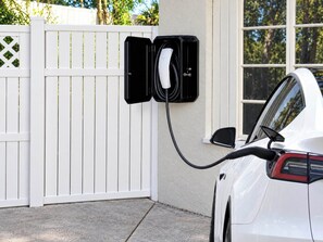 A white car parked next to a white fence with a charging station attached to the fence.