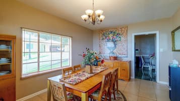 A dining area with a wooden table, chairs, a chandelier, and a window with blinds.