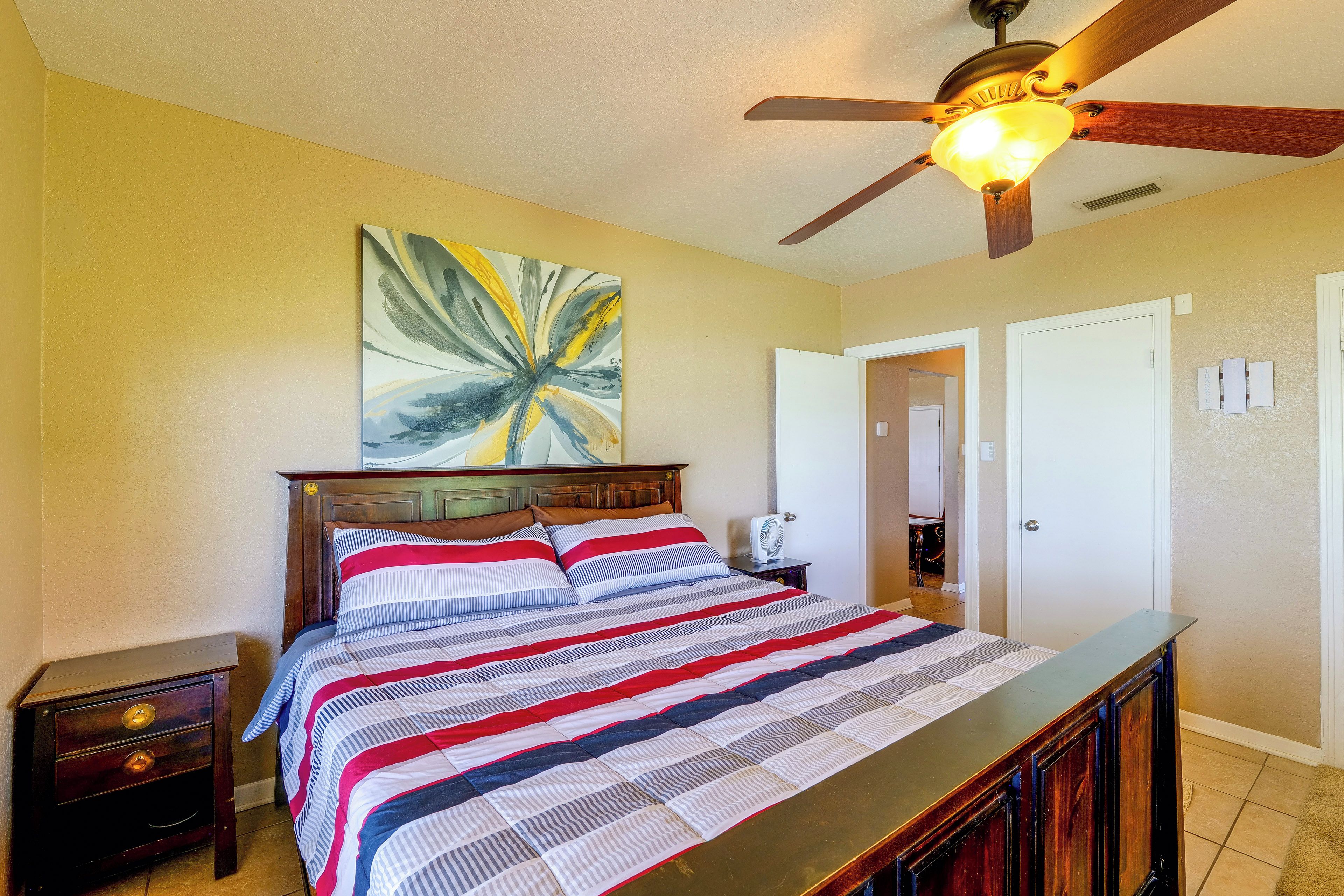 A bedroom with a bed, a ceiling fan, a painting, and a door.