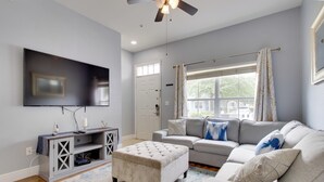A living room with a grey sofa, a white ottoman, a TV on the wall, and a window with curtains.