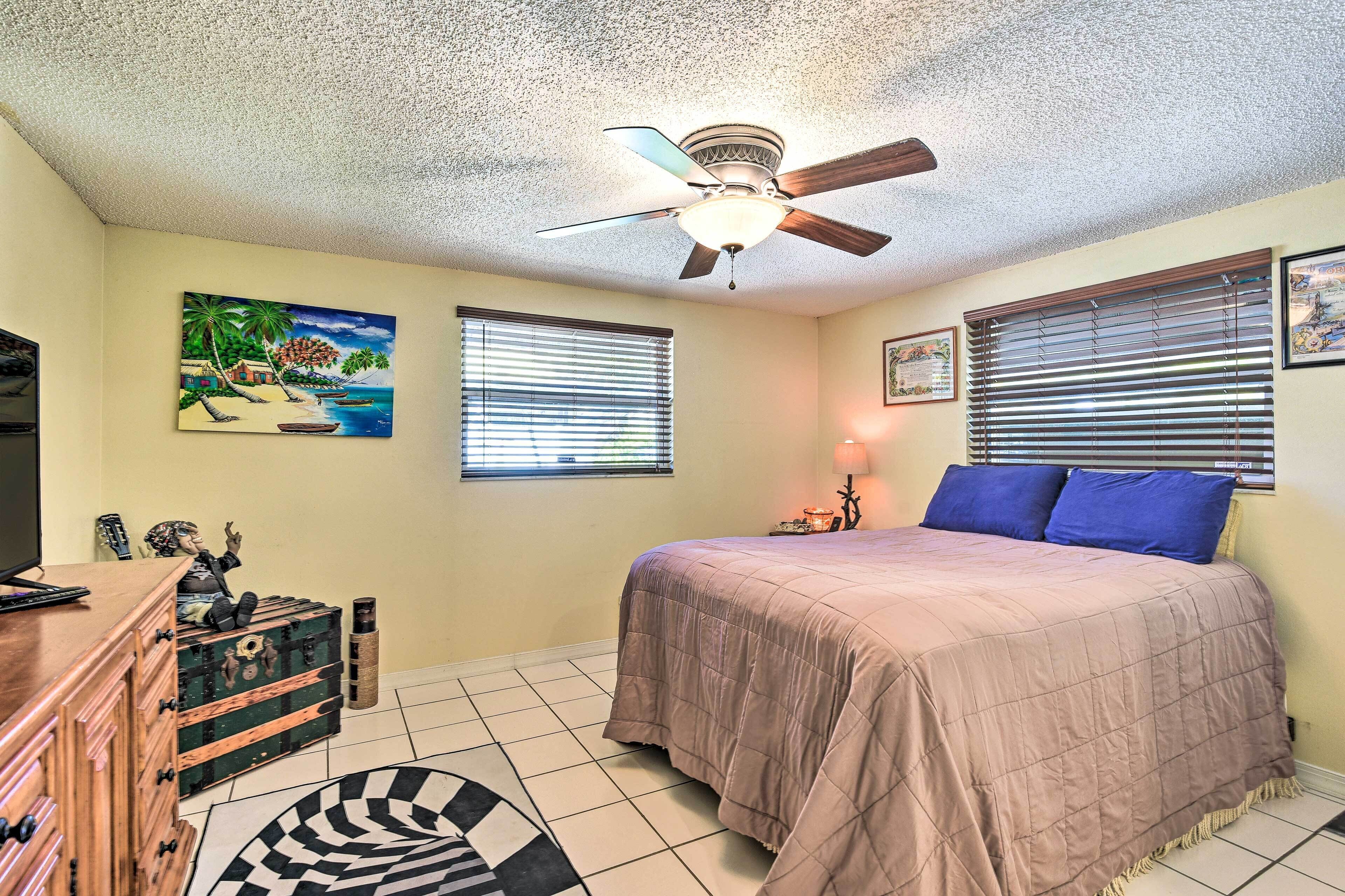 A bedroom with a bed, a ceiling fan, a dresser, a window with blinds, and a painting on the wall.