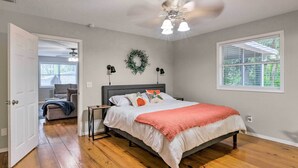 A bedroom with a bed, a nightstand, a wall-mounted lamp, a ceiling fan, a window with blinds, and a door leading to another room.