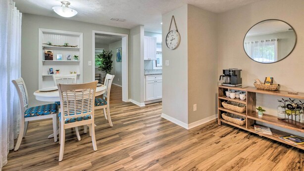 A dining area with a wooden table and chairs, a kitchen in the background, and a shelf with kitchenware.