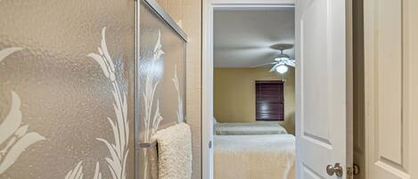 A bathroom with a shower, a towel rack, and a door leading to a bedroom with a bed and a ceiling fan.
