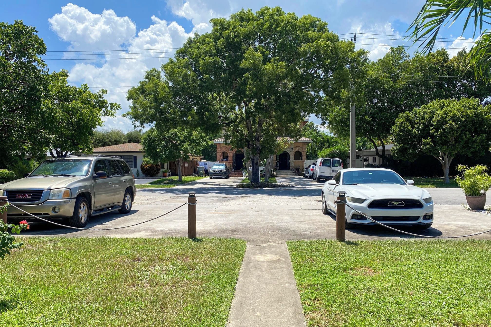 A gated driveway with parked vehicles, a pathway leading to a building, and lush greenery.