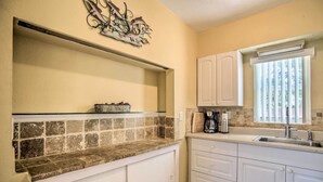Una cocina con armarios blancos, salpicadero de azulejos, cafetera y ventana con persianas.