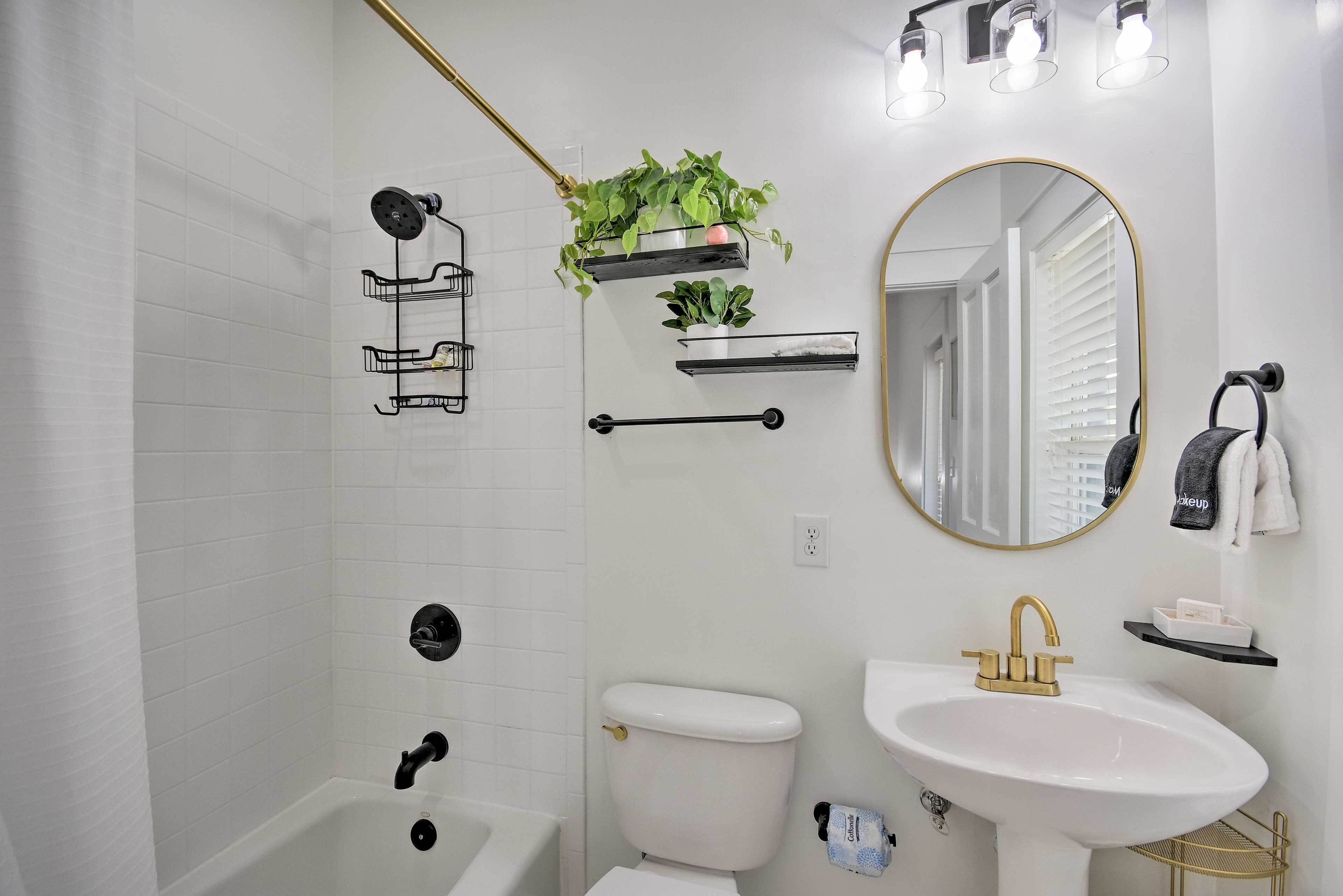 A bathroom with a white bathtub, a white sink, a toilet, and a shower with a gold showerhead. There are plants on the wall and a mirror above the sink.