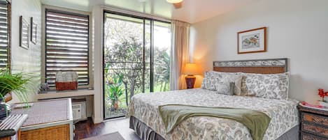 A bedroom with a large bed, a ceiling fan, a window with blinds, a bedside table with a lamp, and a view of greenery outside.