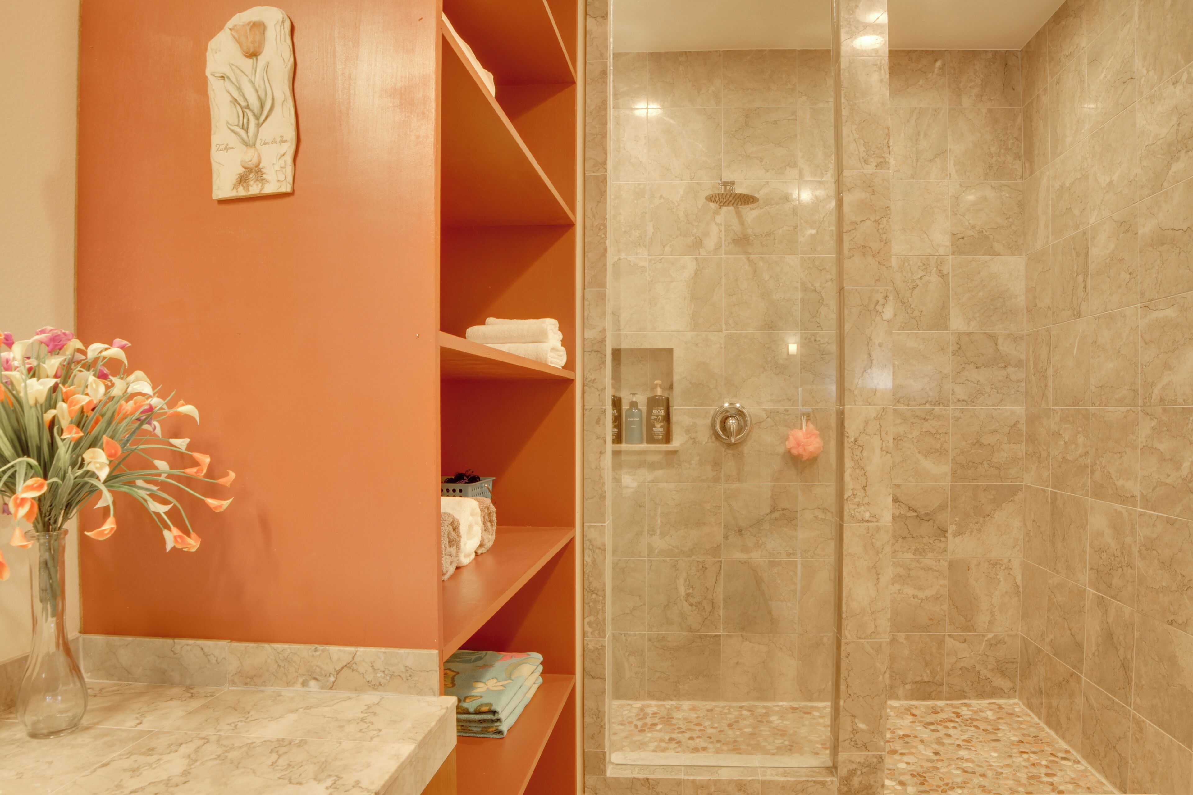 A bathroom with a shower, marble tiles, a vase with flowers, and a shelf with towels.
