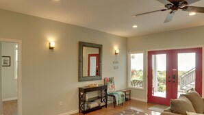 A spacious living room with a red door, a ceiling fan, a console table, and a large window. - Ocean Views Peaceful Retreat Volcano & Beaches (Naalehu)