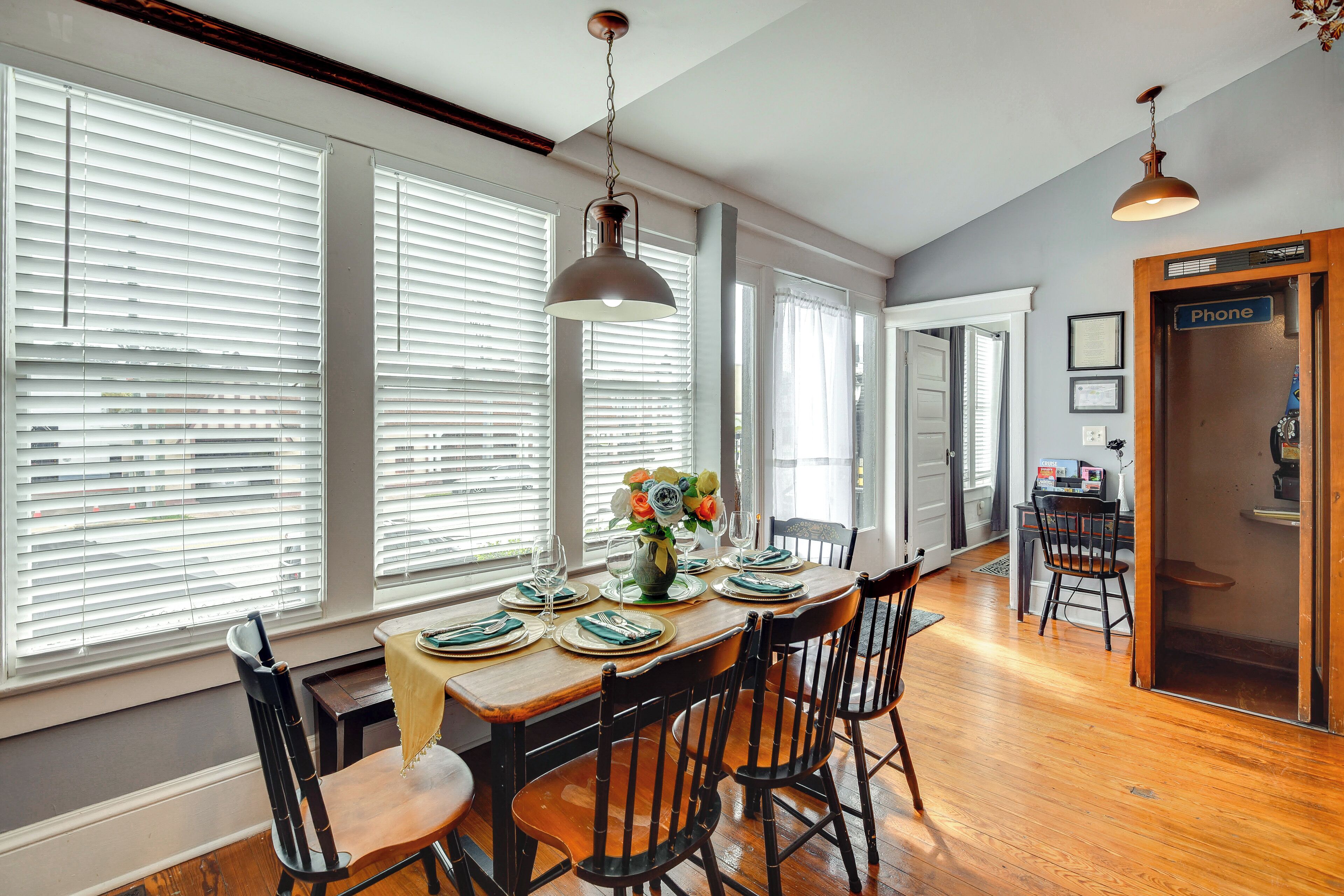 A dining area with a wooden table set for four, flanked by large windows with blinds, a hanging light fixture, and a door labeled 'Phone'.