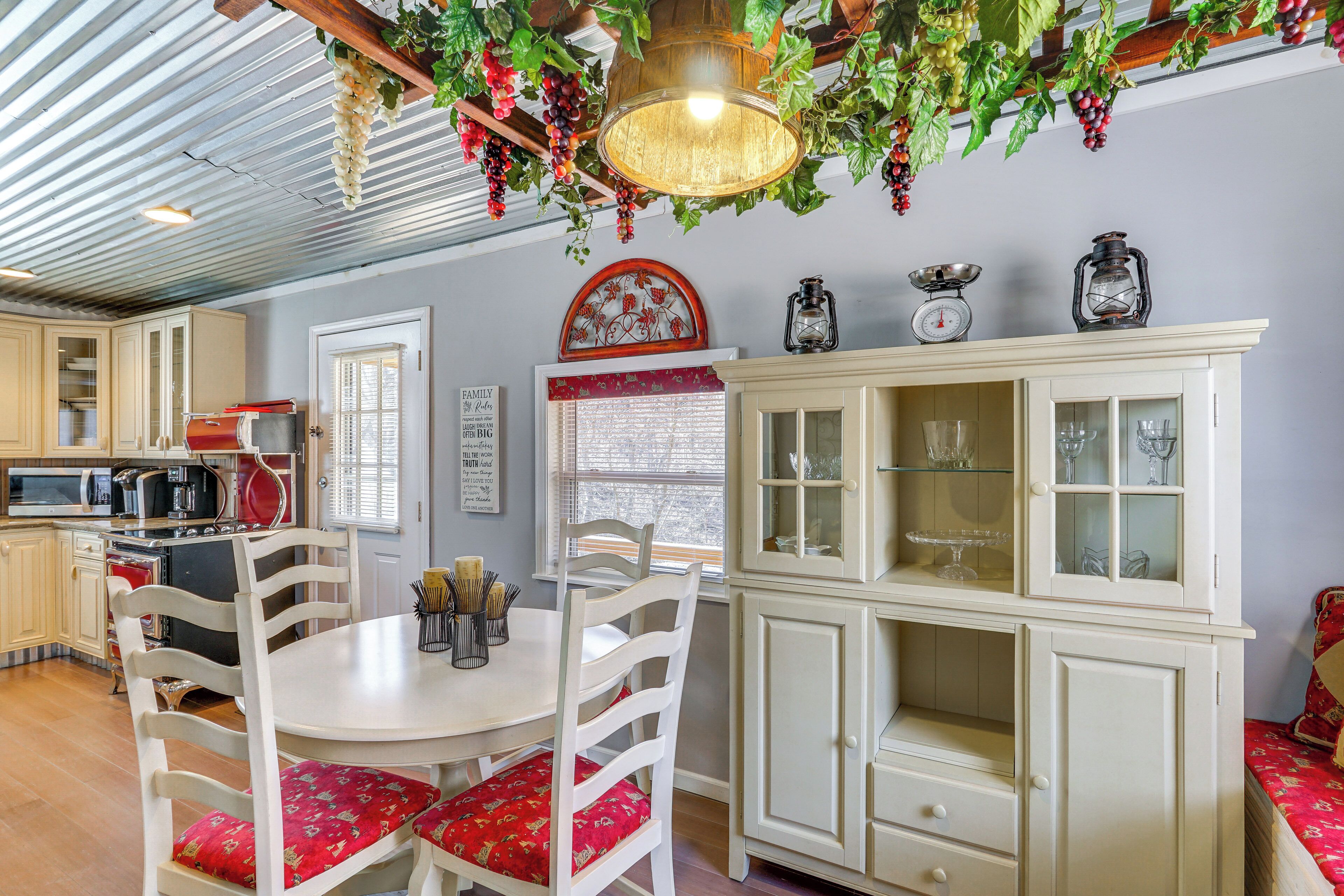 Una cocina con armarios blancos, una mesa de comedor y sillas con cojines rojos. Hay una lámpara colgante decorada con uvas y hojas.