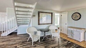 A room with a wooden staircase, two wicker chairs, a small round table, and a white cabinet with a wooden top. - Cozy Waterfront 'island House' on Live Oak Island! (Crawfordville)