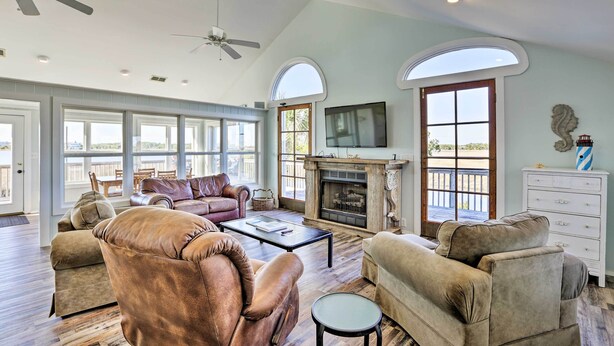 A spacious living room with leather sofas, a fireplace, and large windows offering a view of the outdoors. - Cozy Waterfront 'island House' on Live Oak Island! (Crawfordville)