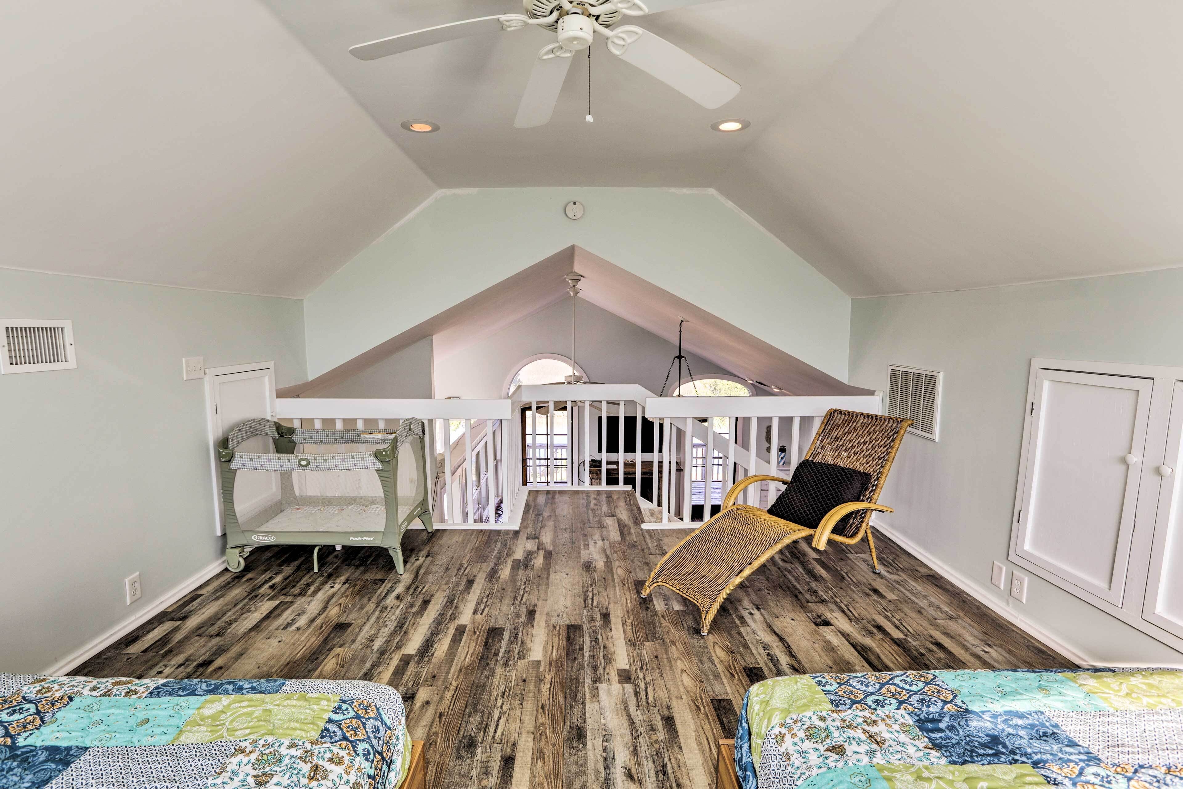 A spacious room with a high ceiling, wooden flooring, a ceiling fan, a crib, a wicker chair, and a bed with a colorful quilt.
