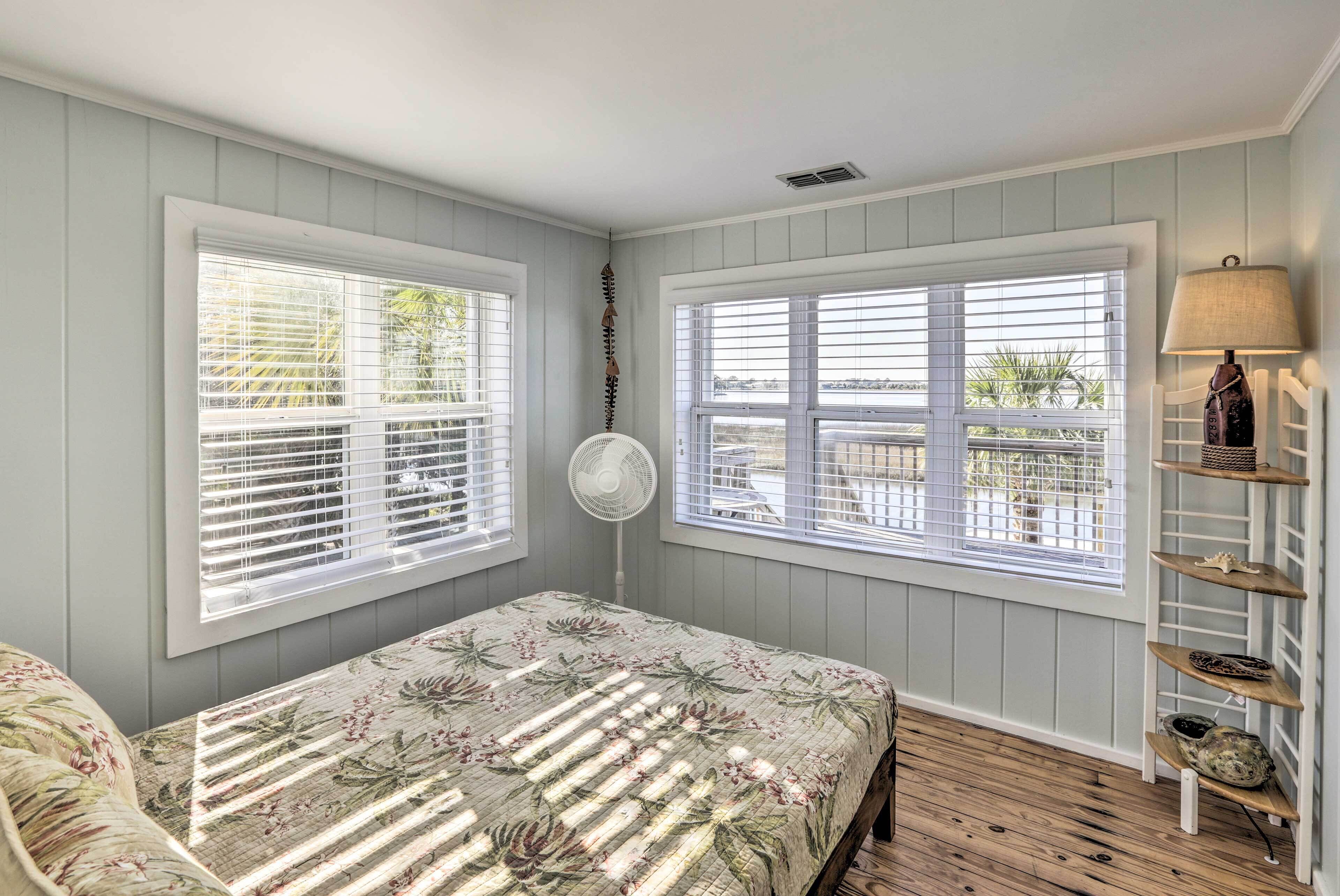A bedroom with two windows, a bed with a floral bedspread, a fan, and a shelf with shoes.