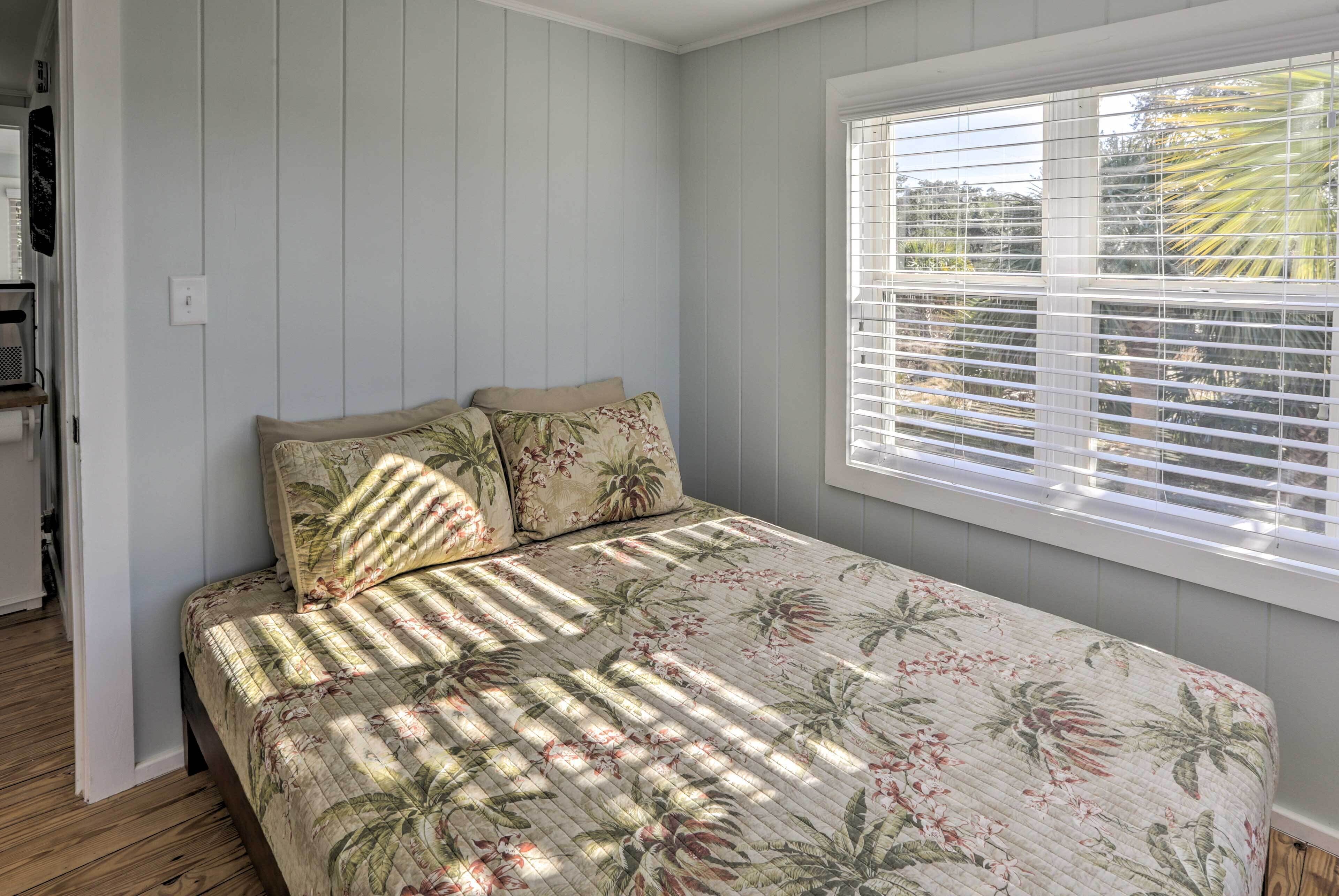 A bedroom with a bed, floral bedding, a window with blinds, and a wooden floor.