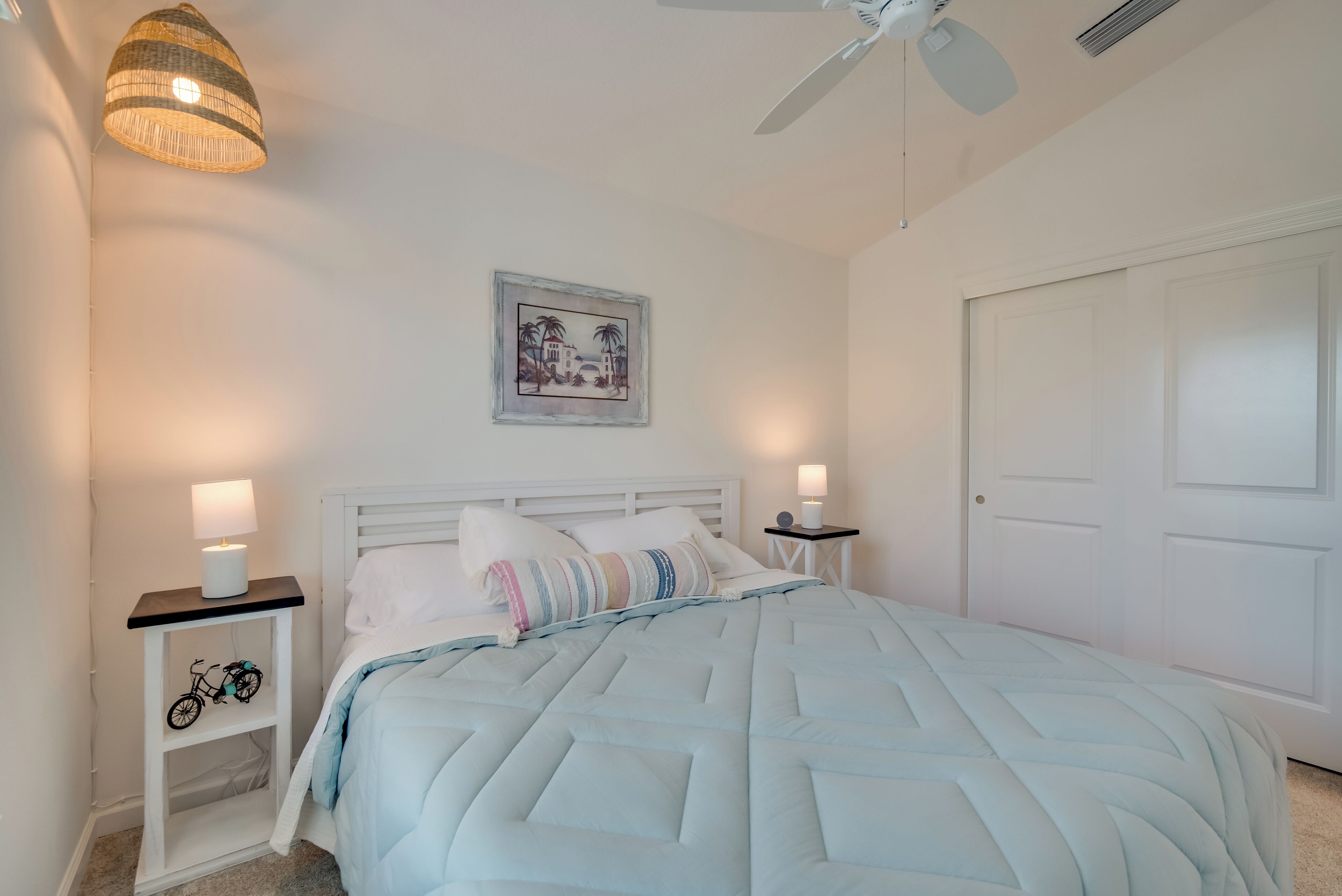 A bedroom with a bed, white walls, a ceiling fan, a framed picture, and bedside lamps.