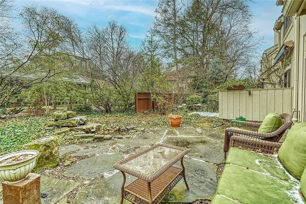 Relax in a serene outdoor space with a wicker table and chairs surrounded by lush greenery and a small wooden shed.