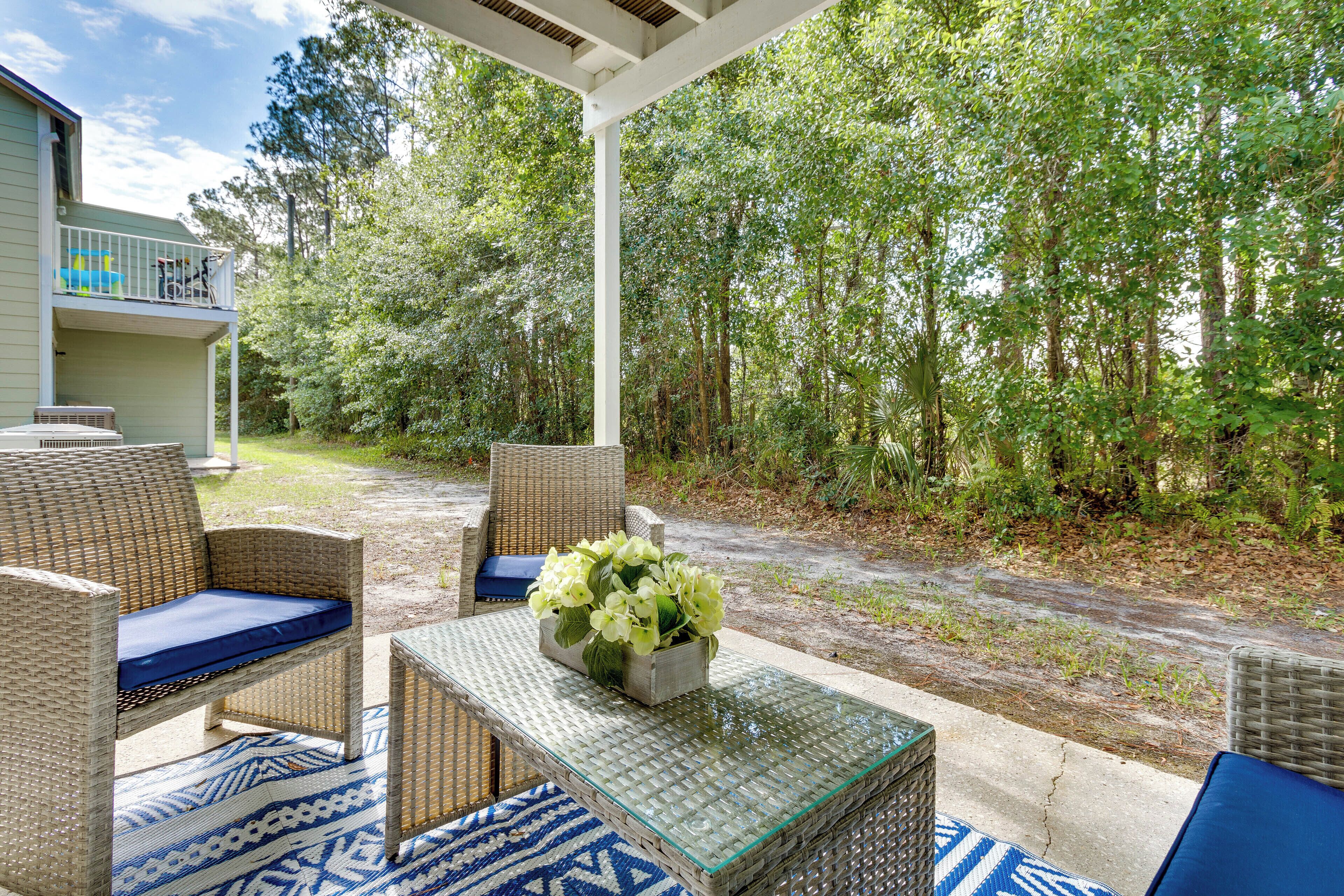 A covered outdoor seating area with wicker furniture and a glass-top table, surrounded by a lush green forest.