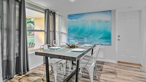 Un coin repas avec une table dressée pour quatre personnes, une grande peinture représentant une vague de l’océan accrochée au mur et, par la fenêtre, une vue sur une maison.