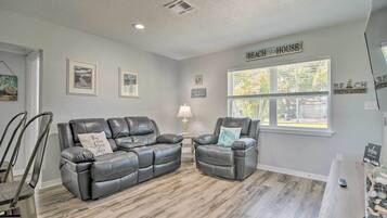 A living room with a gray leather sofa and armchair, a wooden floor, a window with blinds, and a sign that reads "Beach House".