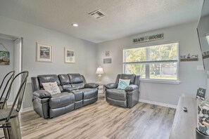A living room with a gray leather sofa and armchair, a wooden floor, a window with blinds, and a sign that reads "Beach House". - Sunny Seminole Home: Grill, Fire Pit & Fenced Yard (Seminole)