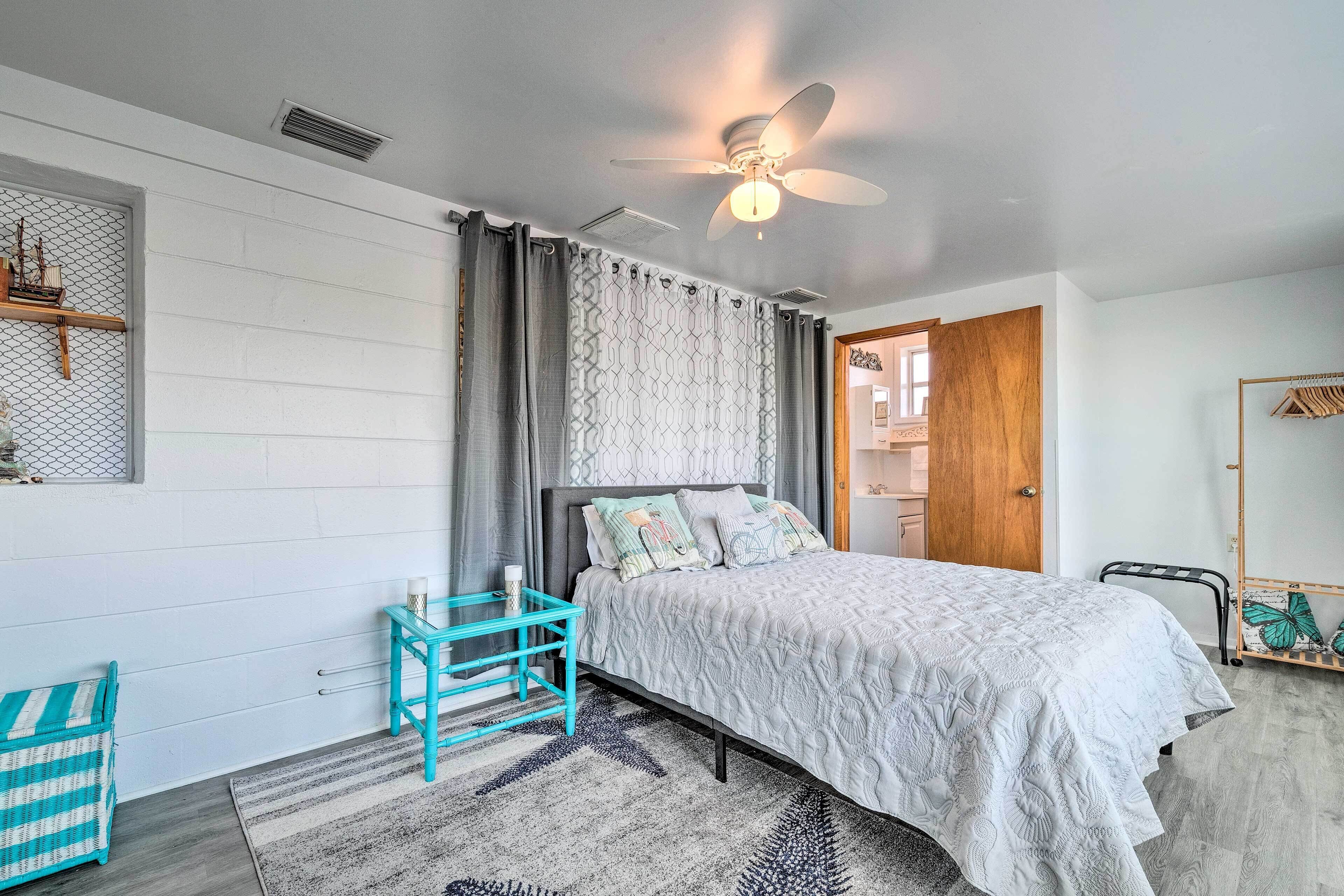 A bedroom with a bed, a ceiling fan, a window with curtains, a small table, and a chair.