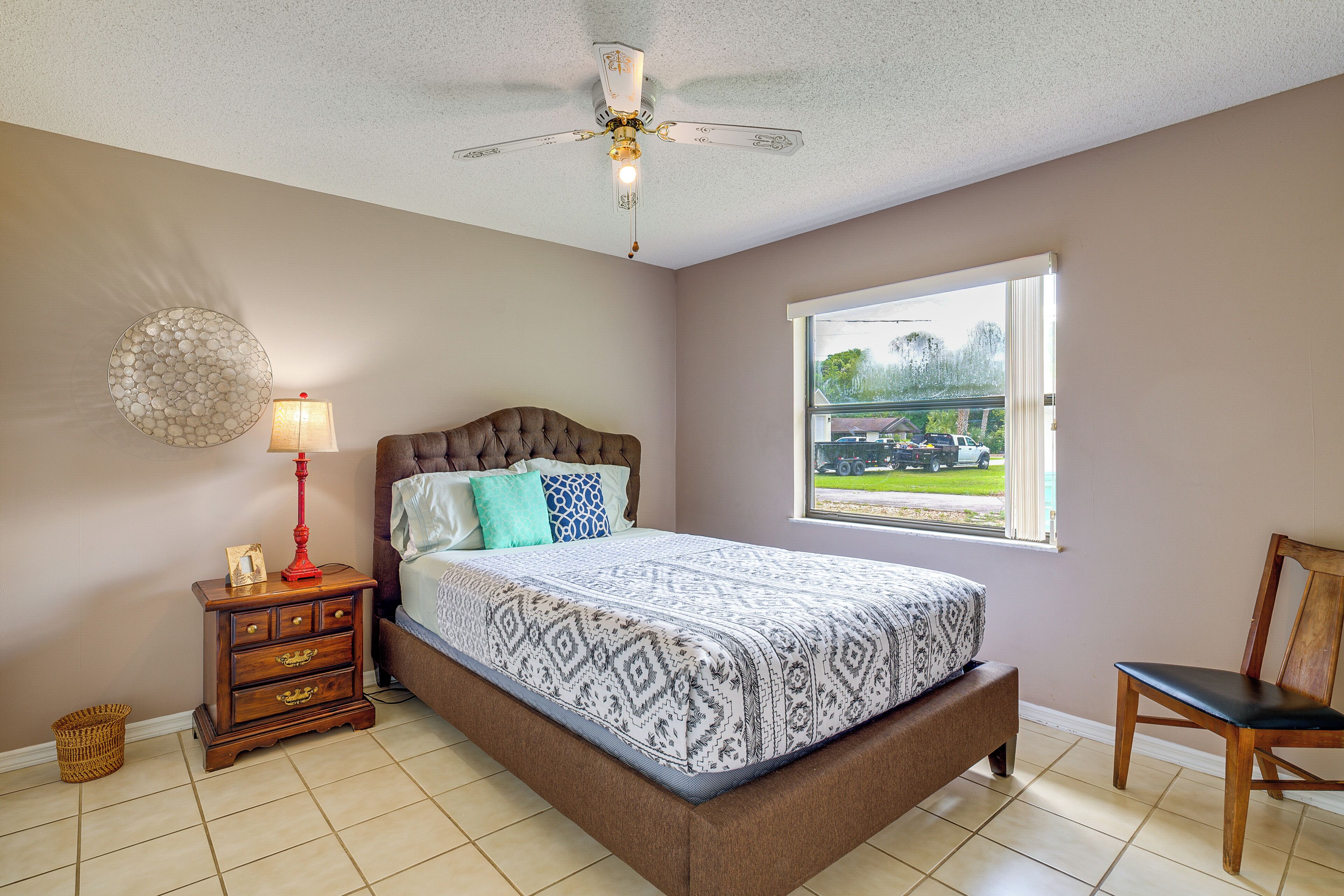 A bedroom with a bed, a nightstand, a lamp, a ceiling fan, a window, and a chair.