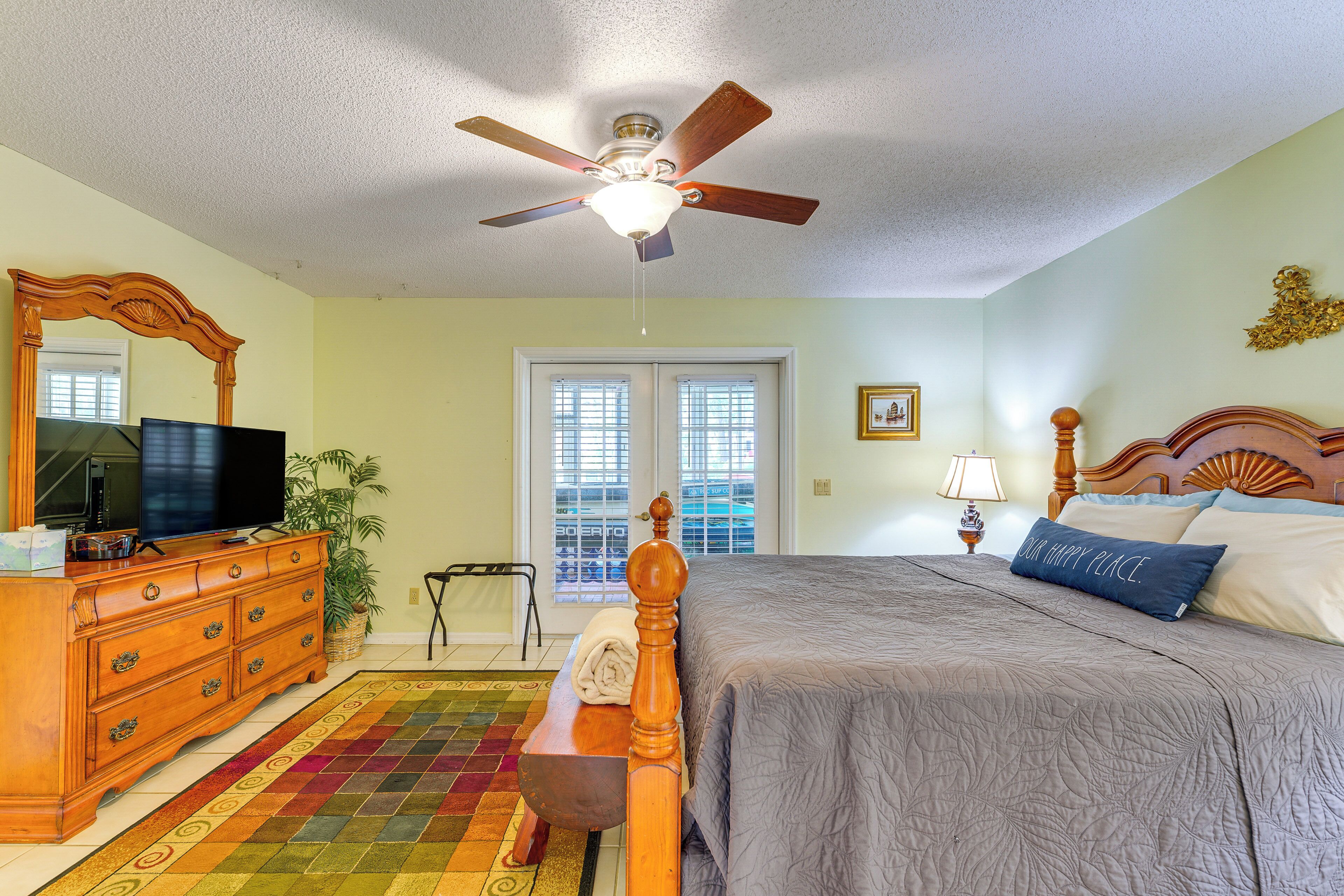 A bedroom with a bed, dresser, television, ceiling fan, windows, and a view of the outdoors.