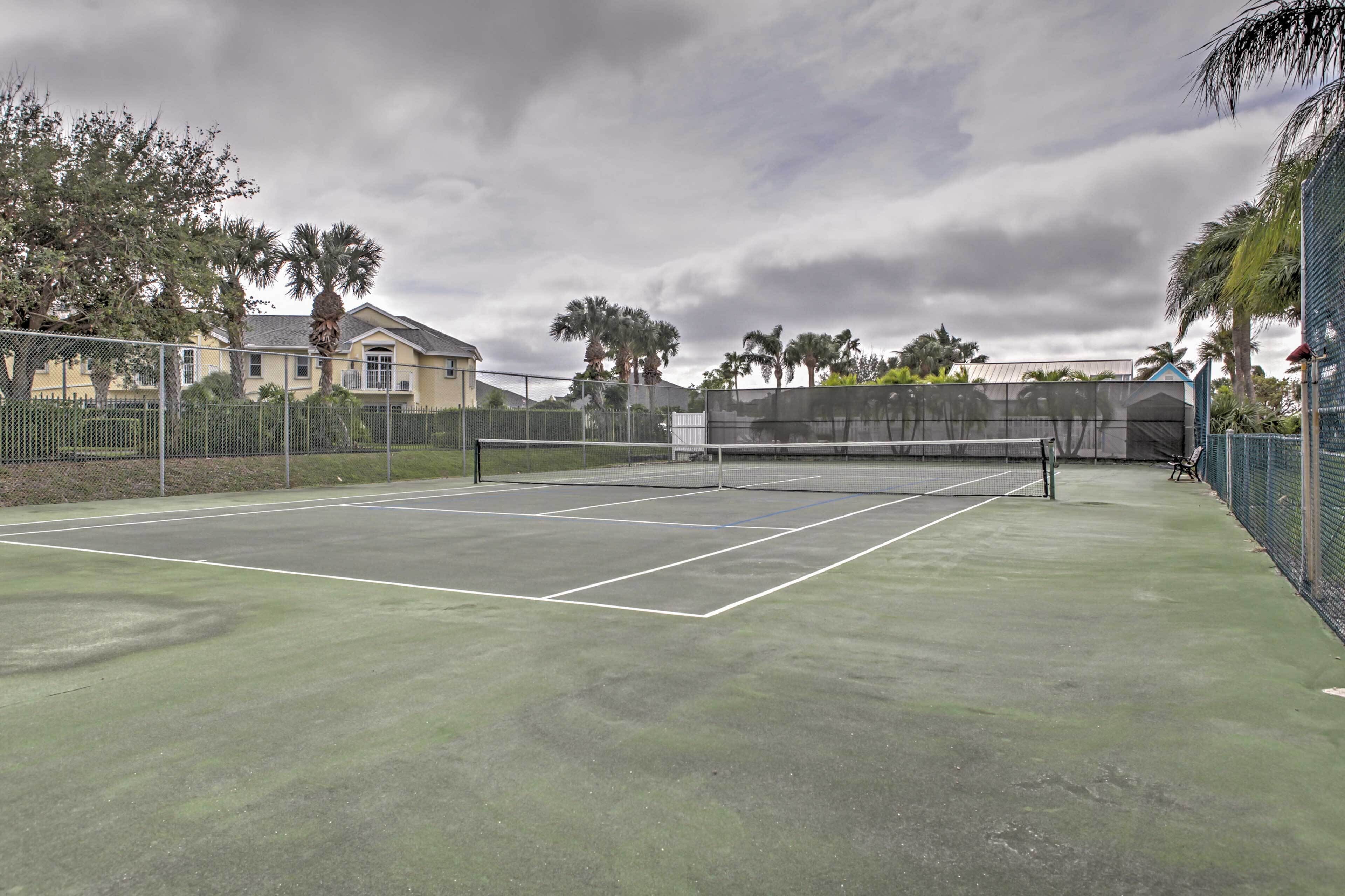 An outdoor tennis court with a net, surrounded by a fence, and palm trees in the background.