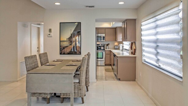 A modern kitchen with stainless steel appliances, a dining area with a long table and wicker chairs, and a large window with blinds. - 'sand Dollar' Jenson Beach Home w/ Pool Access! (Jensen Beach)