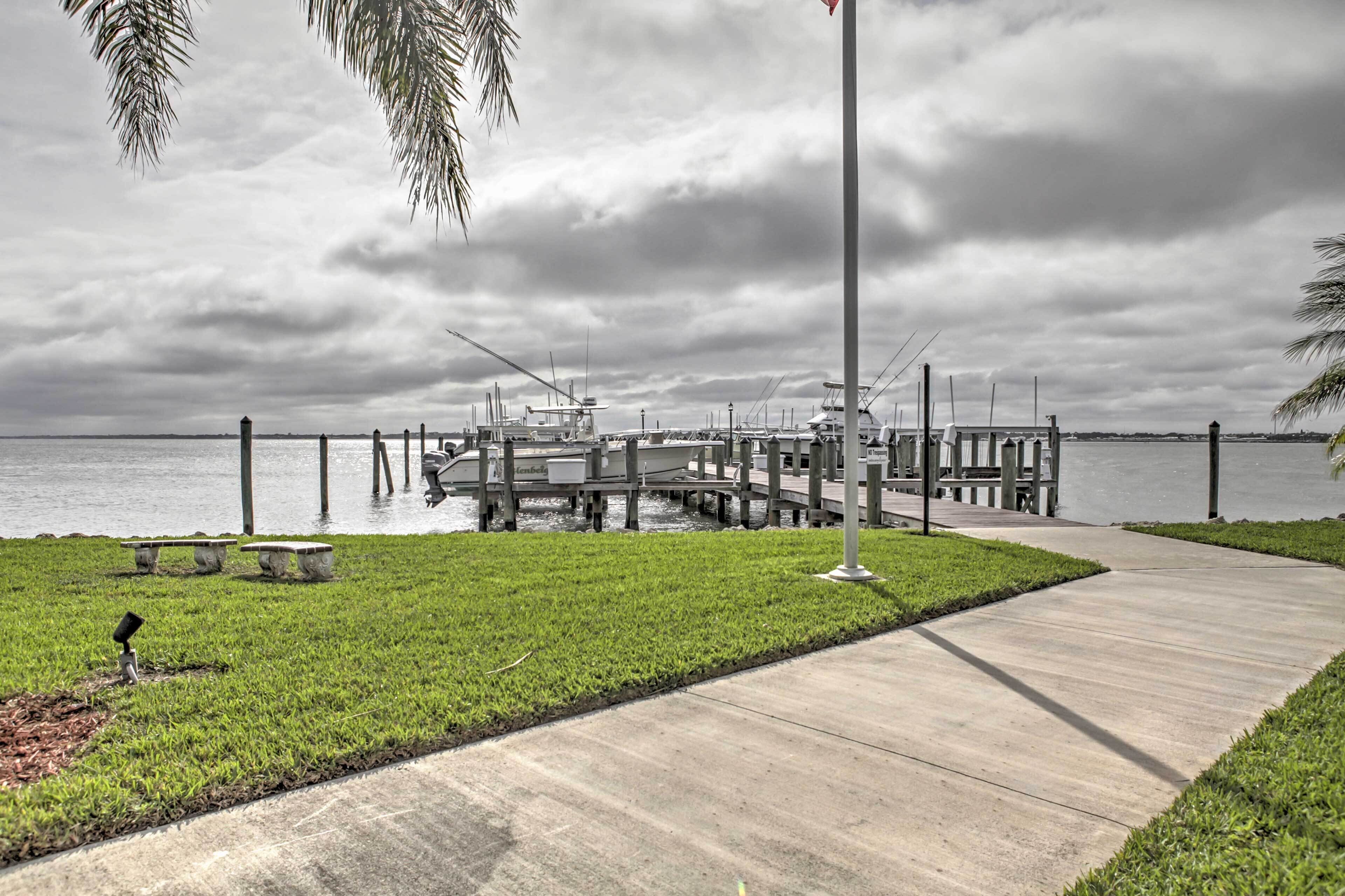 A marina with docks extending into the water, a concrete walkway, and a grassy area with a bench.