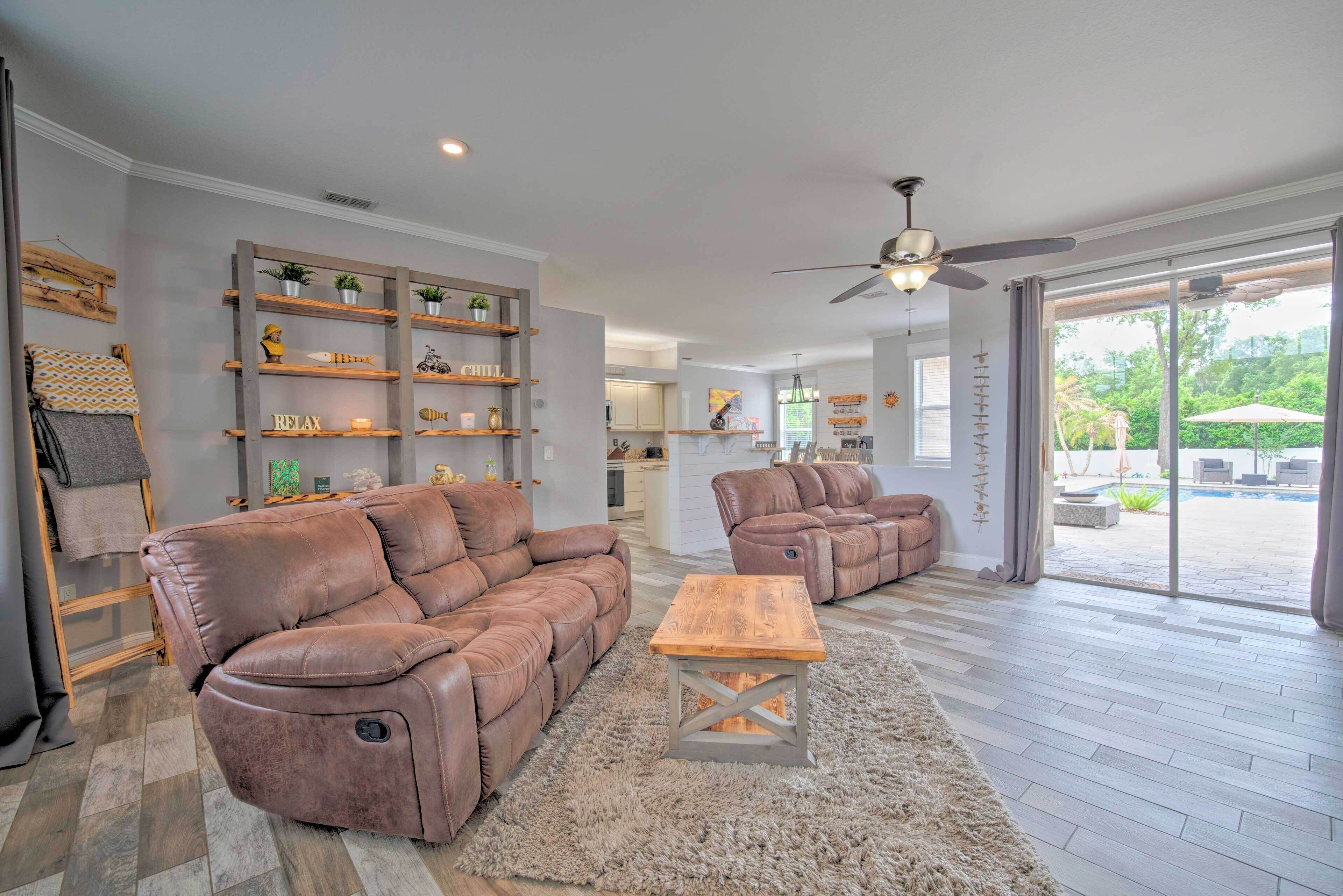 A spacious living room with a brown leather sectional sofa, a wooden coffee table, and a large window offering a view of a pool and greenery.