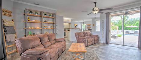 A spacious living room with a brown leather sectional sofa, a wooden coffee table, and a large window offering a view of a pool and greenery.