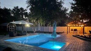 A swimming pool with a tiled deck, surrounded by a fence and trees. There is a covered seating area with string lights.
