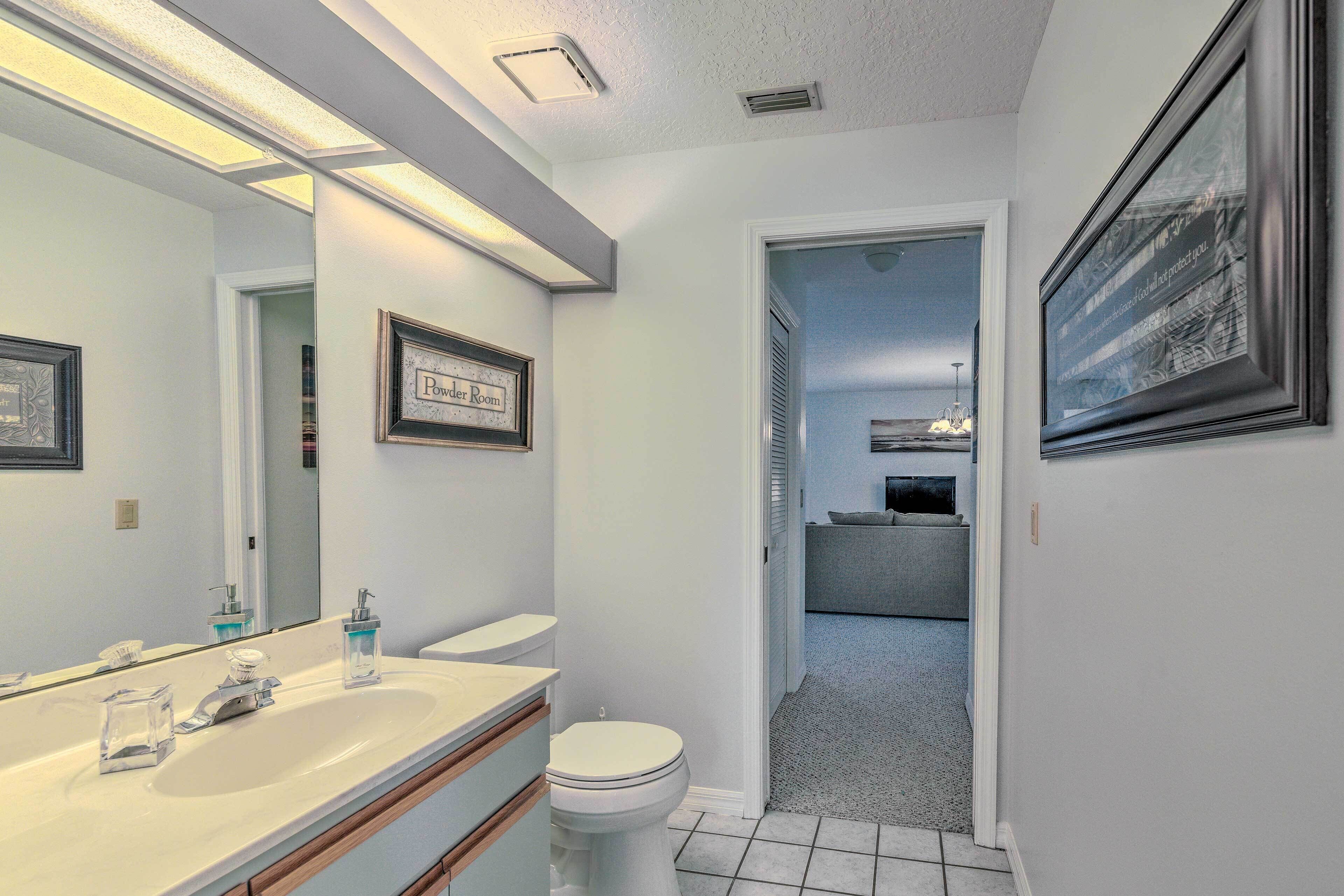 A bathroom with a white sink, a mirror, and a toilet. There is a framed sign on the wall and a framed picture above the mirror.