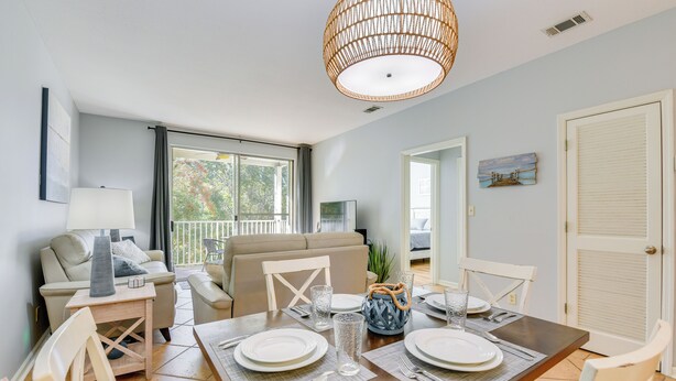 Un coin repas avec une table en bois dressée pour quatre personnes, entourée de chaises blanches. Un lustre en osier est suspendu au-dessus, et une grande fenêtre laisse entrer la lumière naturelle.